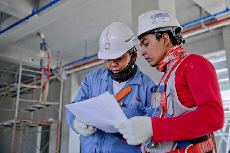 Two contractors in safety helmets reviewing construction plans at a worksite.